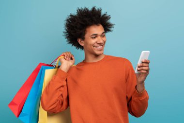 Bir elinde renkli alışveriş poşetleri, diğer elinde akıllı telefonuyla gülümseyen kıvırcık saçlı Afro-Amerikan bir adam..