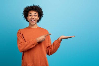 Turuncu kazaklı, neşeli bir Afro-Amerikan adam boş elini gülümseyerek göstererek mekanı kopyalayarak coşku gösteriyor.