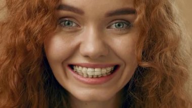 Canlı kıvırcık saçlı genç bir kadın geniş bir gülümsemeyle neşeyi ifade eder. İç mekan ortamında temiz diş tellerini sergiliyor, neşeli tavırlarını vurguluyor..