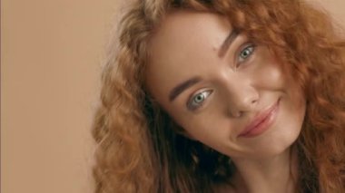 Kıvırcık kızıl saçlı ve çarpıcı hatları olan genç bir kadın kameraya dikkatle bakıyor, eşsiz güzelliğini yumuşak, nötr bir arkaplana karşı sergiliyor..