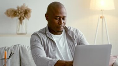 30 'lu yaşlarda bir adam, parlak, modern bir oturma odasında, dizüstü bilgisayarına odaklanmış bir kanepede oturuyor. Yumuşak ışıklandırma ve dekoratif bitkiler üretkenlik için rahat bir atmosfer yaratır..