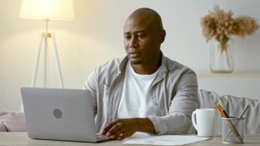 Modern bir ev ofisinin ahşap masasında oturan bir adam dizüstü bilgisayarıyla meşgul oluyor. Odaklanmış görünüyor, hafif ışık etrafındaki sakin atmosferi güçlendiriyor..