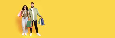 Alışverişe. Kredi kartı ve alışveriş poşetleriyle gülümseyen orta doğulu çift, stüdyoda sarı arka planda sezonluk satışlar sırasında bir sürü alışveriş satın almış. Tam boy çekim