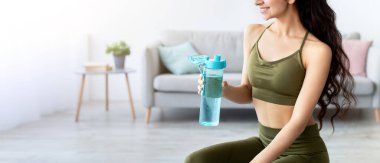 Spor kıyafetli çekici Hintli bayan evde bir şişe temiz suyla fitness topunun üzerinde oturuyor. Çekici Asyalı kadın ev içi eğitim sırasında susuz kalıyor, içeride çalışıyor.