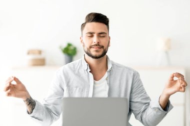 Sakin olun. Erkek serbest çalışan biri dizüstü bilgisayarda çalışırken, gözleri kapalı oturup iş yerinde stres atmak için meditasyon yapıyor. Meditasyon ve rahatlama egzersizi konsepti