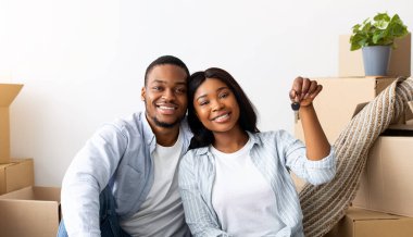 Ev anahtarı ve karton kutuları olan mutlu Afro-Amerikan eşleri kameraya bakıyor ve gülümsüyor, mülklerinde yerde oturuyorlar. Genç siyahi ev sahipleri mutlu mesut kendi evlerine taşınıyorlar.