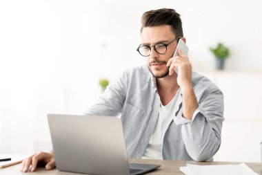 Akıllı telefondan konuşan yakışıklı bir genç, müşteriyle konuşan, iş yerinde dizüstü bilgisayarla çalışan. Girişimci iş görüşmesi yapıyor. İletişim Kavramı