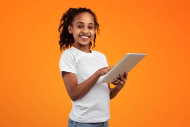 Dijital Yerliler, Çocuklar İçin Uygulama. Güzel gülümseyen Afro-Amerikan genç kız, dijital tableti turuncu stüdyo arka planında tutuyor ve kullanıyor. Kameraya bakıyor ve poz veriyor.