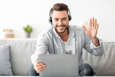 Uzaktan iletişim konsepti. Kulaklıklı, dizüstü bilgisayara el sallayan, koltukta otururken video görüşmesi yapan mutlu bir milenyum adamı. İnternette ailesiyle veya arkadaşlarıyla sohbet eden genç bir adam.