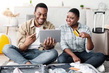 Neşeli Afrikalı turistler dijital tablet ve kredi kartıyla online turne rezervasyonu yapıyorlar, internete bakıyorlar ve evlerinde birlikte seyahat biletleri alıyorlar. Seyahat Hizmetleri ve Uygulamalar Konsepti