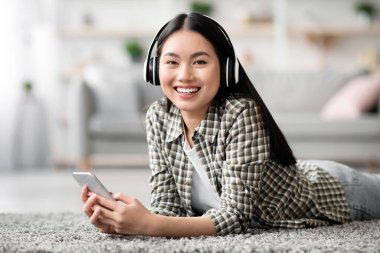 Evde yerde yatarken kablosuz kulaklık ve akıllı telefon kullanarak müzik dinleyen Asyalı rahat bir bayan. Mutlu genç bir kadın cep telefonuyla eğleniyor.
