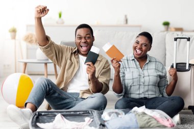 Seyahat biletleriyle poz veren, bavullarıyla evde yaz tatiline hazır ve kameraya gülümseyen mutlu siyah çift. Uzun zamandır Beklenen Tatili Kutlayan Turistler