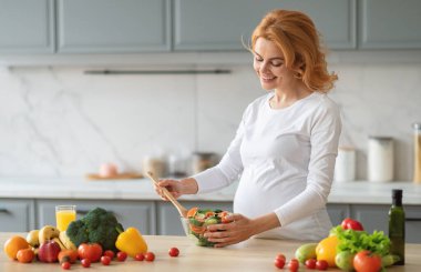 Hamileliğindeki bir kadın, taze sebze ve meyvelerle çevrili şık bir mutfakta renkli bir salatayı neşeyle karıştırıyor. Sıcacık bir ortamda sağlıklı bir yemek hazırlanıyor..