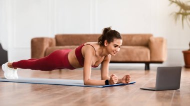 Yurt İçi Spor Konsepti. Gülümseyen Sporcu Kadın Oturma odasında, Plank pozisyonunda dizüstü bilgisayar ekranına bakıyor, bilgisayar başında alıştırma yapıyor. Çevrimiçi Çalıştırma