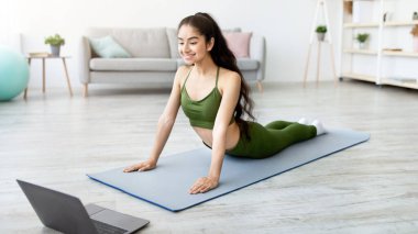 Tam boy Hintli kadın kobra asana yapıyor. Evde dizüstü bilgisayardan yoga videosu eğitimi alıyor. Koronavirüs kilitlenmesi sırasında sağlıklı omurga için ev içi antrenman yapan sağlıklı Doğulu kadın.