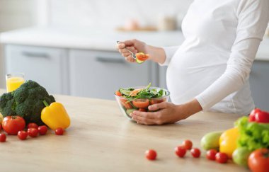 Hamile bir kadın, parlak bir mutfakta sağlıklı bir salata hazırlamaktan hoşlanır. Domates, brokoli ve dolma biber gibi taze sebzeler sanatsal olarak düzenlenir..