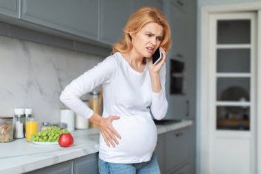 Hamile bir kadın, iyi ışıklandırılmış bir mutfakta, karnını tutarken gözle görülür şekilde rahatsız olur. Telefonda, muhtemelen durumu hakkında konuşuyor, yakınlarda meyvelerle..