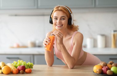 Kulaklıklı bir kadın elinde bir bardak portakal suyu tutarken gülümsüyor. Taze meyveler mutfak tezgahına yayılarak canlı ve sağlıklı bir atmosfer yaratıyor..