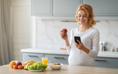 Hamile bir kadın bir elinde elma tutarken diğer elinde modern mutfağında dururken telefonunu kontrol eder. Taze meyve ve bir bardak meyve suyu yakında, fotokopi odası.