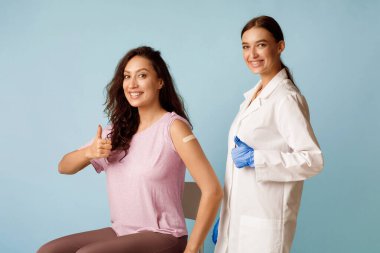 Kadın hemşire ve aşılanmış hasta bayan jestleri, mavi arkaplan üzerinde Covid-19 aşı kampanyasını onaylıyor. Coronavirus Önleme Konsepti. Stüdyo Çekimi