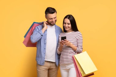Genç bir çift birbirine yakın duruyor, akıllı telefona bakarken neşeli bir anı paylaşıyorlar. Renkli alışveriş çantaları taşıyorlar, eğlenceli günlerini sergiliyorlar..