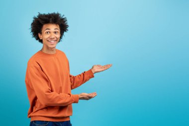 Turuncu kazak giyen genç bir Afro-Amerikalı adam sanki eğlenceli bir şey sunuyormuş gibi gülümsüyor ve kollarını uzatıyor. Parlak mavi arkaplan pozitif titreşim ekler, boşluğu kopyala