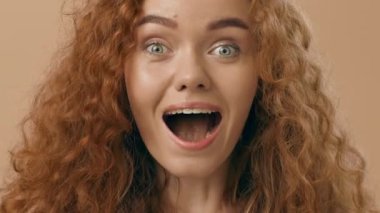 Kıvırcık kızıl saçlı genç bir kadın şaşkın görünüyor, geniş gözleri ve açık ağzı şaşkınlık ve zevki yansıtıyor. Işık arka planı onun canlı ifadesini arttırıyor..