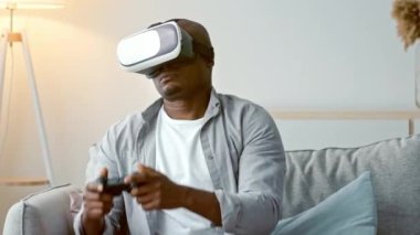 Genç bir Afrikalı Amerikalı adam şık bir oturma odasında rahat bir kanepede otururken sanal gerçeklik kulaklığı takıyor. Odaklanmış ve oyun anının tadını çıkarıyor..