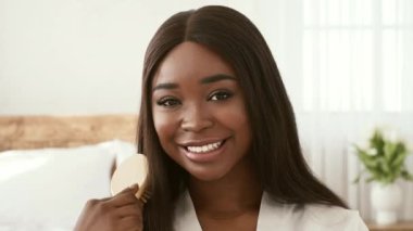 Uzun, pürüzsüz saçları olan neşeli bir Afro-Amerikalı kadın rahat, aydınlık bir alanda oturur, tahta bir fırça tutar ve izleyiciye içten bir şekilde gülümser. Rahatlamış ve mutlu görünüyor..