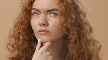 Çarpıcı kıvırcık saçlı genç bir kadın derin düşüncelere dalmış, çenesini elinin üzerine koyuyor. İnce bir arkaplana karşı ciddi bir ifadeyle yukarı bakıyor..