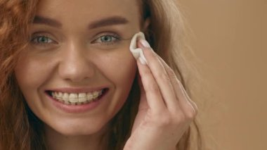 Genç bir kadın, banyoda pamuk pedi ile yüz temizleyiciyi uygularken parlak bir şekilde gülümsüyor. Sabah güneşi onun neşeli yüz ifadesini ve canlı kıvırcıklarını vurguluyor..