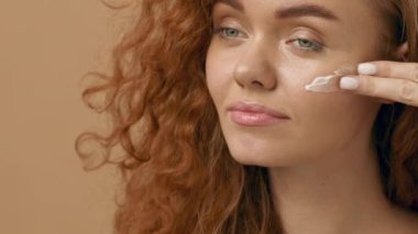 Kıvırcık saçlı neşeli genç bir kadın evde otururken yüzüne nemlendirici sürüyor. Sıcak arka plan, onun ışıl ışıl gülümsemesini ve izlediği cilt rutinini arttırıyor..