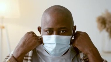 30 'lu yaşlarda Afro-Amerikalı bir adam maskesini rahat bir ev ortamında ayarlıyor. Atmosfer sakin ve zorlu zamanlarda kişisel güvenliğin altını çiziyor..
