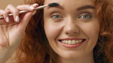 Kıvırcık kızıl saçlı genç bir kadın kaş fırçası kullanırken gülümsüyor. Parlak iç güzelliği, onun neşeli yüz ifadesini geliştirir. Rahat bir ortamda güzellik rutinini gözler önüne serer..