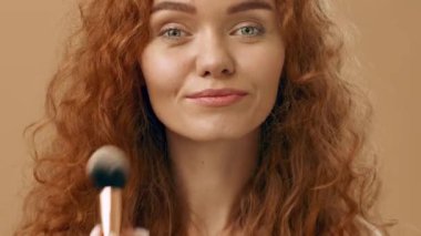 Neşeli, kıvırcık, kızıl saçlı genç bir kadın kendine güvenerek ayakta duruyor ve gülümsüyor. Rahatlamış ve cana yakın görünüyor. Tarafsız bir bej arka planda duruyor. Sıcak bir ortam yaratıyor..