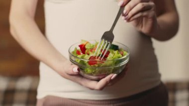 Hamile bir kadın bir kase taze salata, çatal elinde, sıcak evinde dinlenirken sağlıklı sebzelerin ve sebzelerin tadını çıkarıyor..
