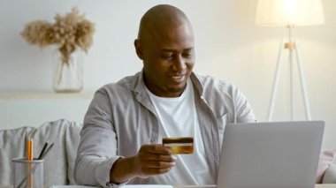 30 'lu yaşlarında bir adam evde dizüstü bilgisayarının önünde kredi kartı tutarken gülümsüyor. Çevrimiçi alışverişle meşgul. Oturduğu yerde rahat bir anın tadını çıkarıyor..