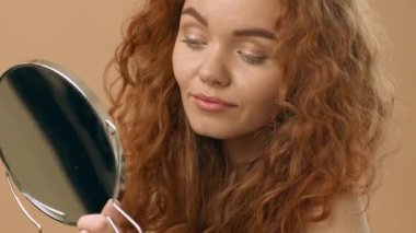 Kıvırcık saçlı bir kadın, parmaklarını nazikçe saçlarında gezdiren bir aynaya bakıyor. Sıcak tonlu arka plan onun rahatlamış ifadesini ve kişisel anını tamamlıyor..