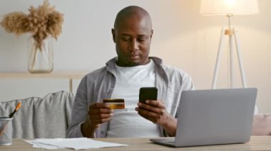 30 'lu yaşlarda bir adam, elinde kredi kartı tutarken akıllı telefonunu kullanarak modern bir evde rahatça oturabilir. Ekonomisini rahat bir atmosferde yönetmeye odaklanmış durumda..