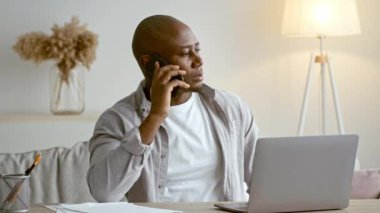 30 'lu yaşlarda Afro-Amerikan bir adam ev ofisinde otururken bir telefon görüşmesi yapıyor. Önünde bir dizüstü bilgisayar ve masanın üzerine dağılmış kağıtlarla odaklanmış durumda..