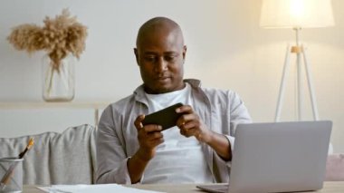Afro-Amerikalı adam evde rahatlıyor, akıllı telefonuna odaklanıyor ve yakınlarda dizüstü bilgisayarı olan bir masada rahatça oturuyor. Doğal ışık konforlu boşluğu doldurur.