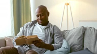 30 'lu yaşlarda bir adam, parlak ve modern bir oturma odasında rahat bir kanepede kitap okumaktan zevk alır. Atmosfer sakin, rahatlama ve boş zamanı teşvik ediyor..