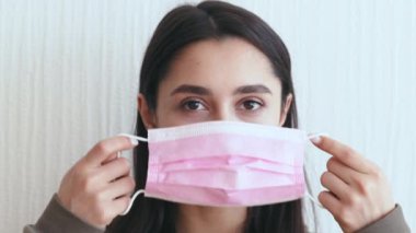 Genç beyaz bir kadın evde otururken pembe bir maske takıyor. Hastalık nedeniyle izolasyon sırasında grip ya da soğuk algınlığı belirtileri gösteriyor..
