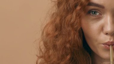 Kıvırcık kızıl saçlı genç bir kadın, sıcak tonlu bir arka planda oturarak pipetten içkisini yudumluyor. Yüzündeki ifade bir rahatlama anı yakaladığını gösteriyor..