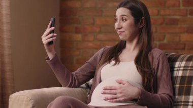 Neşeli hamile bir kadın kanepede oturuyor, karnını sarıyor ve sıcak, rahat bir atmosferle çevrili iken telefonuyla selfie çekiyor..