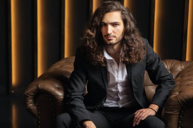 Kendine güvenen, uzun saçlı genç bir adam modern bir ofiste deri bir koltukta zarif bir şekilde oturur. Ilık ışıklandırma onun iş kıyafetini vurgulayarak, davetkar bir şirket ortamı yaratıyor..