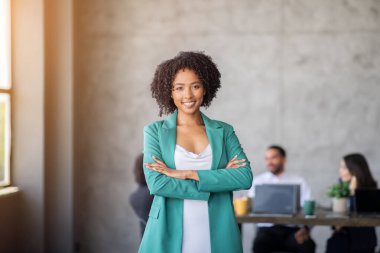 In a contemporary office featuring large windows, a confident woman stands with crossed arms. Behind her, a diverse team engages in discussion by a table, highlighting teamwork and collaboration. clipart