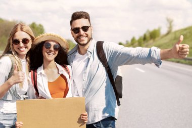Otobanda otostop çeken, el hareketleriyle hareket eden, içi boş tabelalar tutan mutlu arkadaş portresi. Mutlu gençler yazın otostop çekiyorlar.