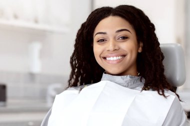 Modern stomatolojik klinikteki randevusu sırasında koltuğunda oturan mutlu, genç Afrikalı Amerikalı kadın dişçi doktorla görüşmeye hazır, kameraya bakıyor ve gülümsüyor.,