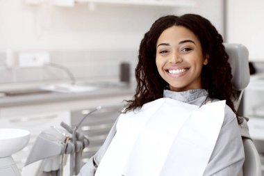 Diş Hizmetleri Konsepti. Stomatoloji kliniğinde sandalyede oturan mutlu, gülümseyen zenci kadın portresi. Afrikalı kadın hasta, dişçi kontrolüne ve tedavisine hazır, kameraya bakıyor.